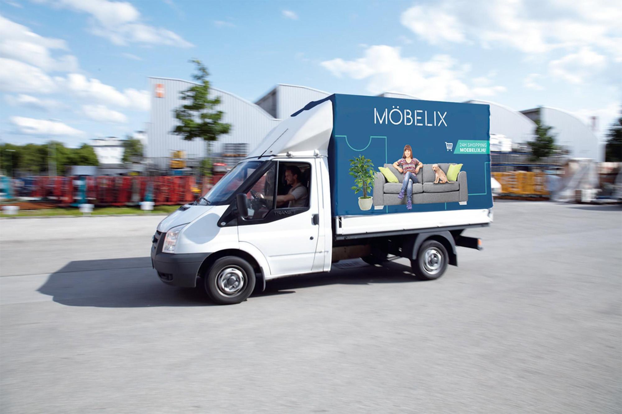 Möbelix feliratú, reklámgrafikával dekorált teherautó halad egy áruházparkolóban, a háttérben ipari épületekkel.
