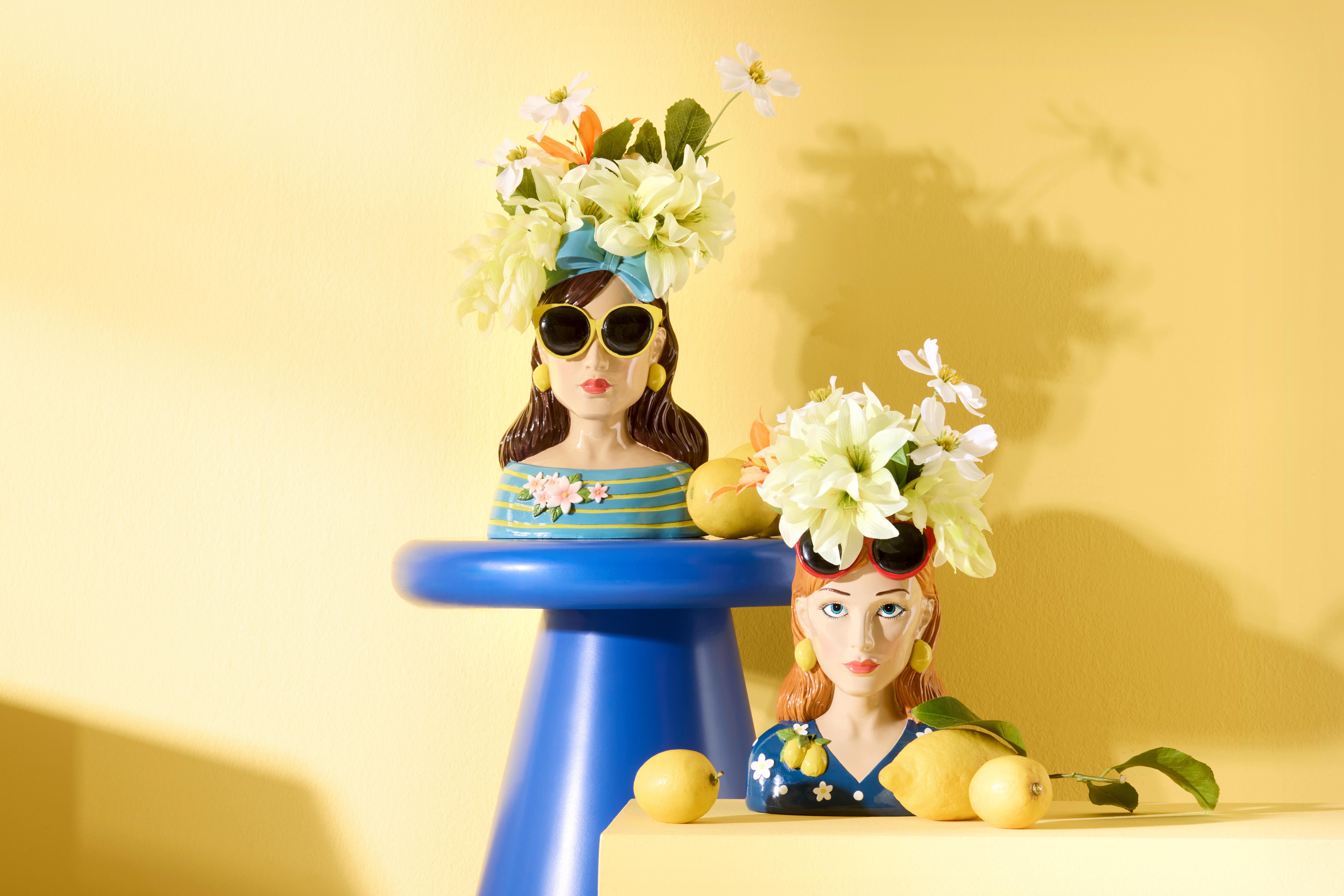 Dvě keramické vázy Zene ve tvaru ženských bust s květinami a slunečními brýlemi na modrém bočním stolku Marino, doplněné citony na žlutém pozadí.