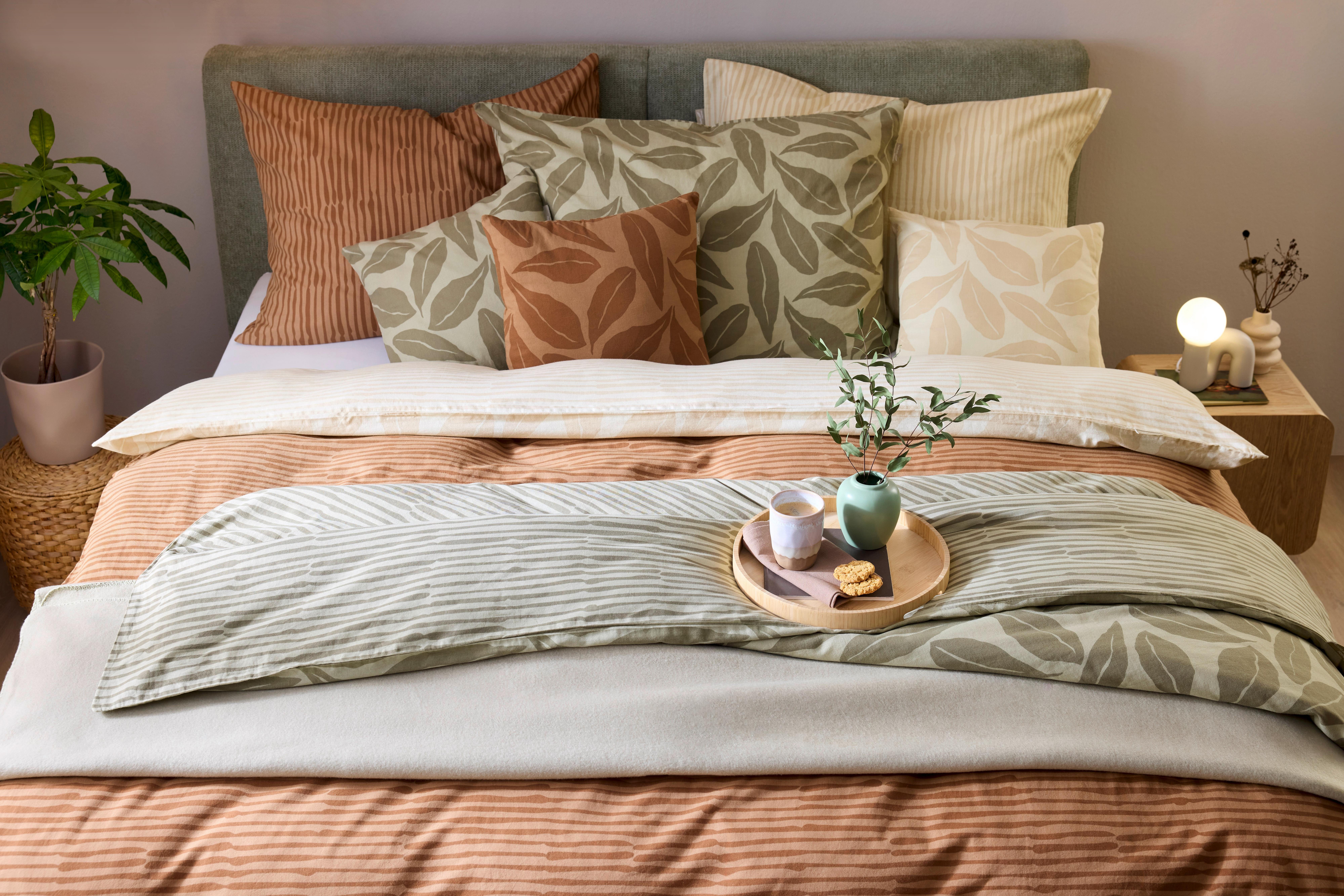 Spálňa v štýle Urban Nature s vrstvenými obliečkami v zemitých tónoch a vankúšmi s listovým motívom. Atmosféru dopĺňa drevený stolík, jemné svetlo a prírodné dekorácie.
