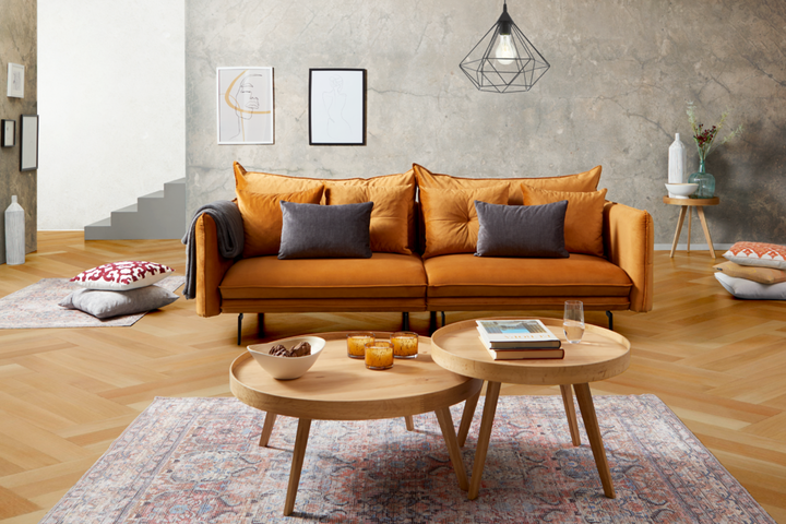 Vor einer Wand in Beton-Optik steht ein oranges Sofa mit runden Beistelltischen auf einem roten Orientteppich.