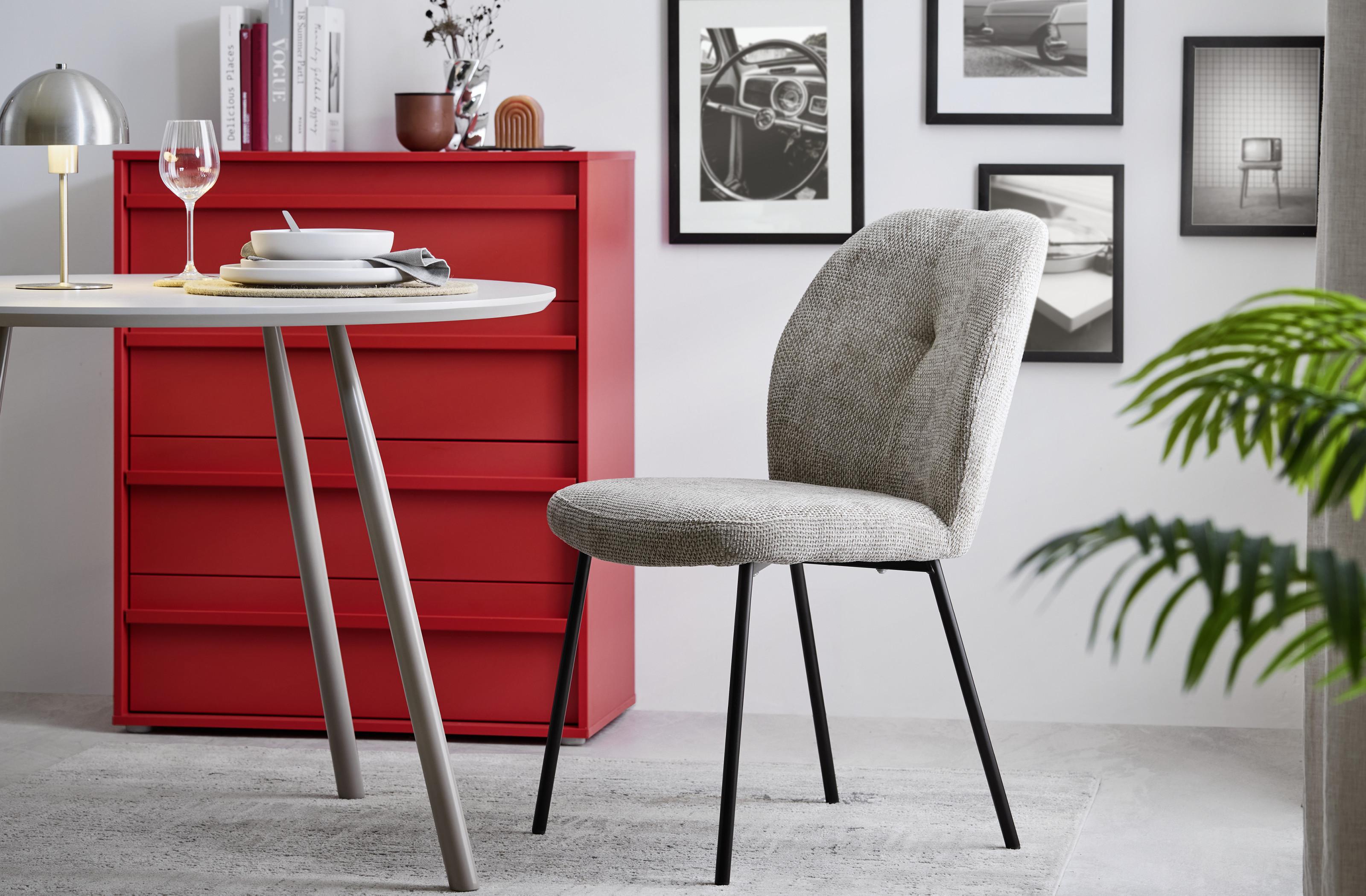 Svetlá jedáleň v retro štýle so sivou čalúnenou stoličkou, okrúhlym stolom a výraznou červenou komodou doplnenou dekoráciami a čiernobielymi obrazmi.