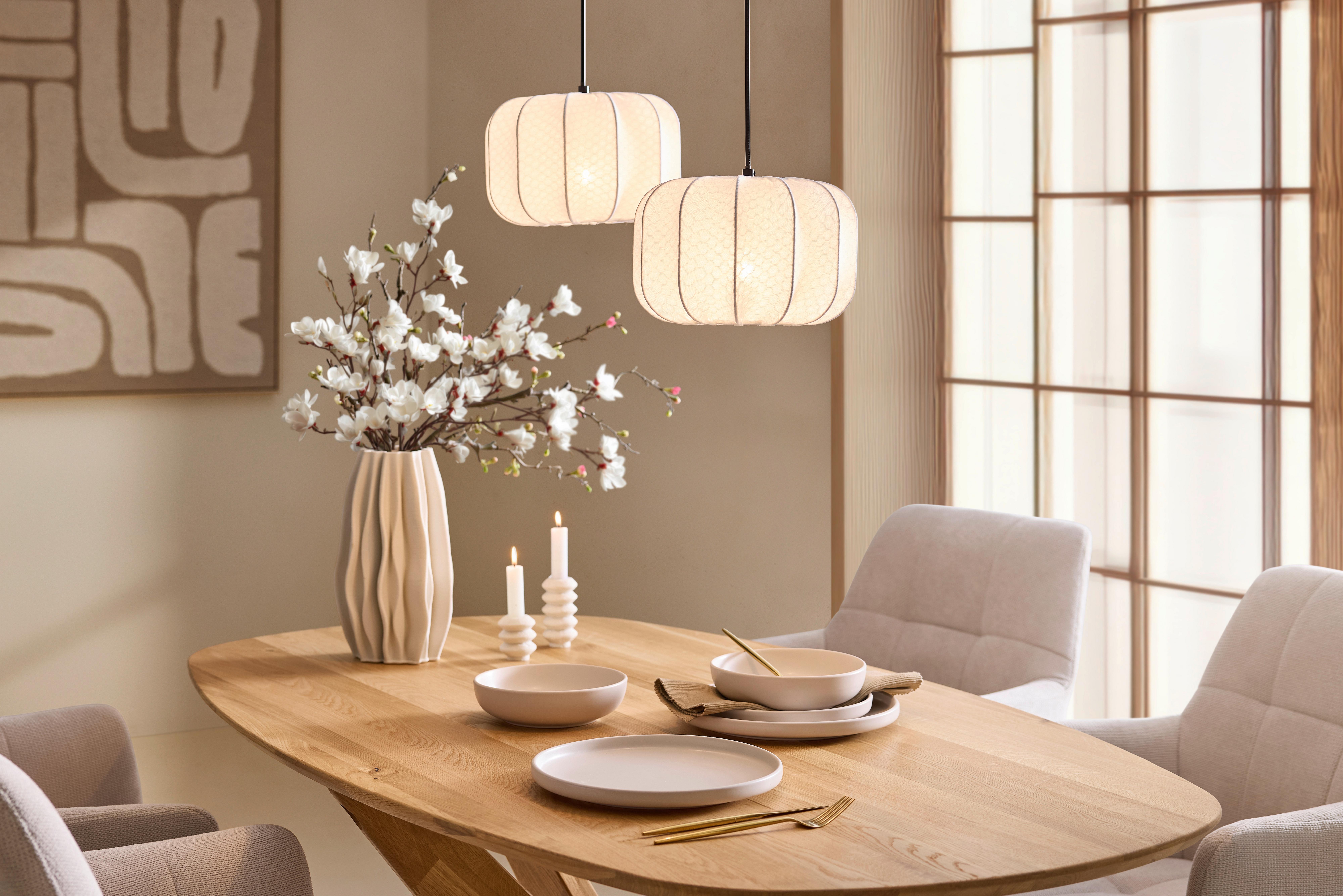 Thought Prestretý drevený jedálenský stôl s minimalistickým riadom a vázou s kvetmi, osvetlený dvoma závesnými lampami.
