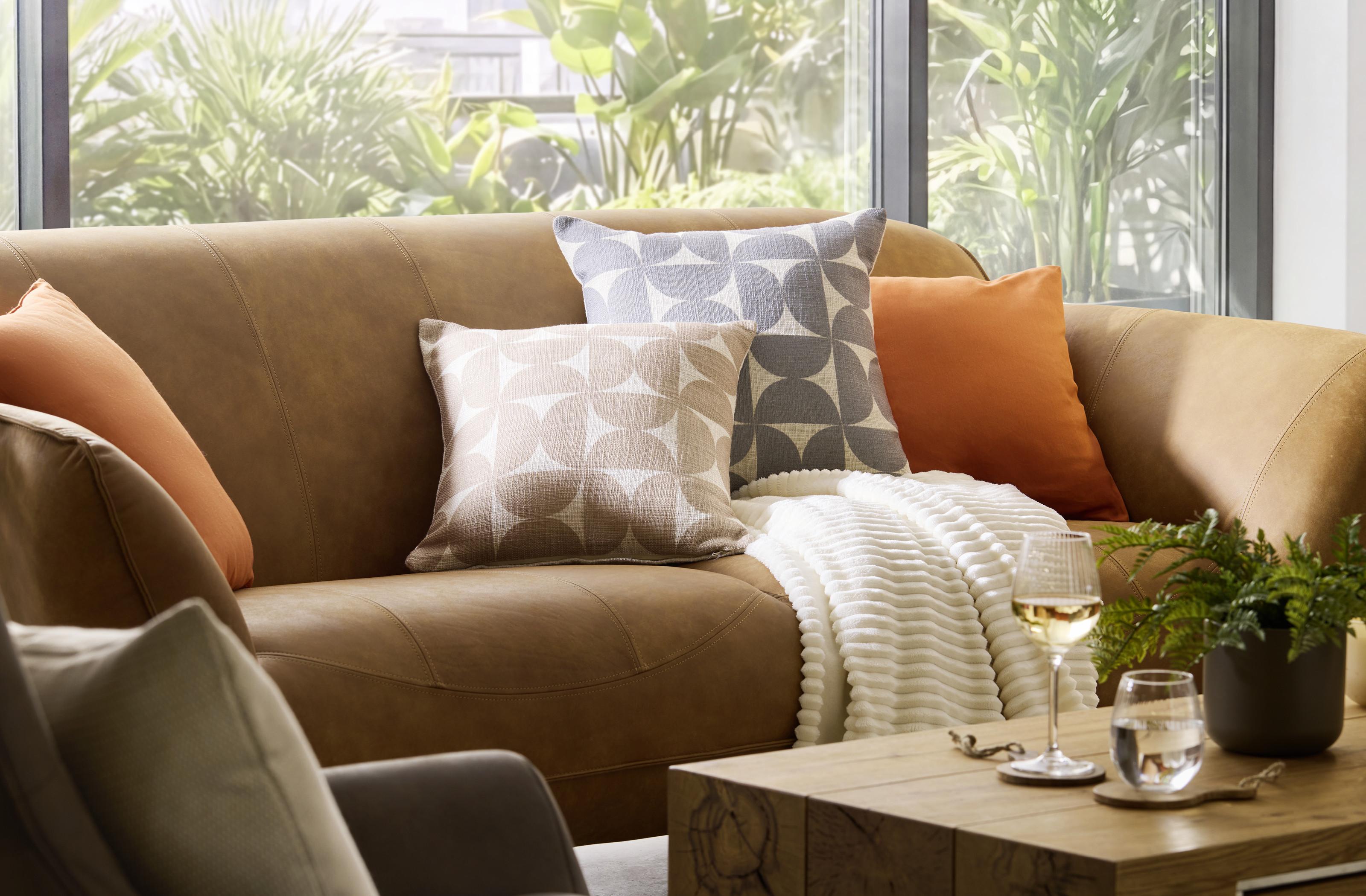 Retro obývačka s hnedou sedačkou, vzorovanými vankúšmi, jemnou dekou a prírodnými doplnkami ktoré vytvárajú útulný a štýlový relaxačný priestor.