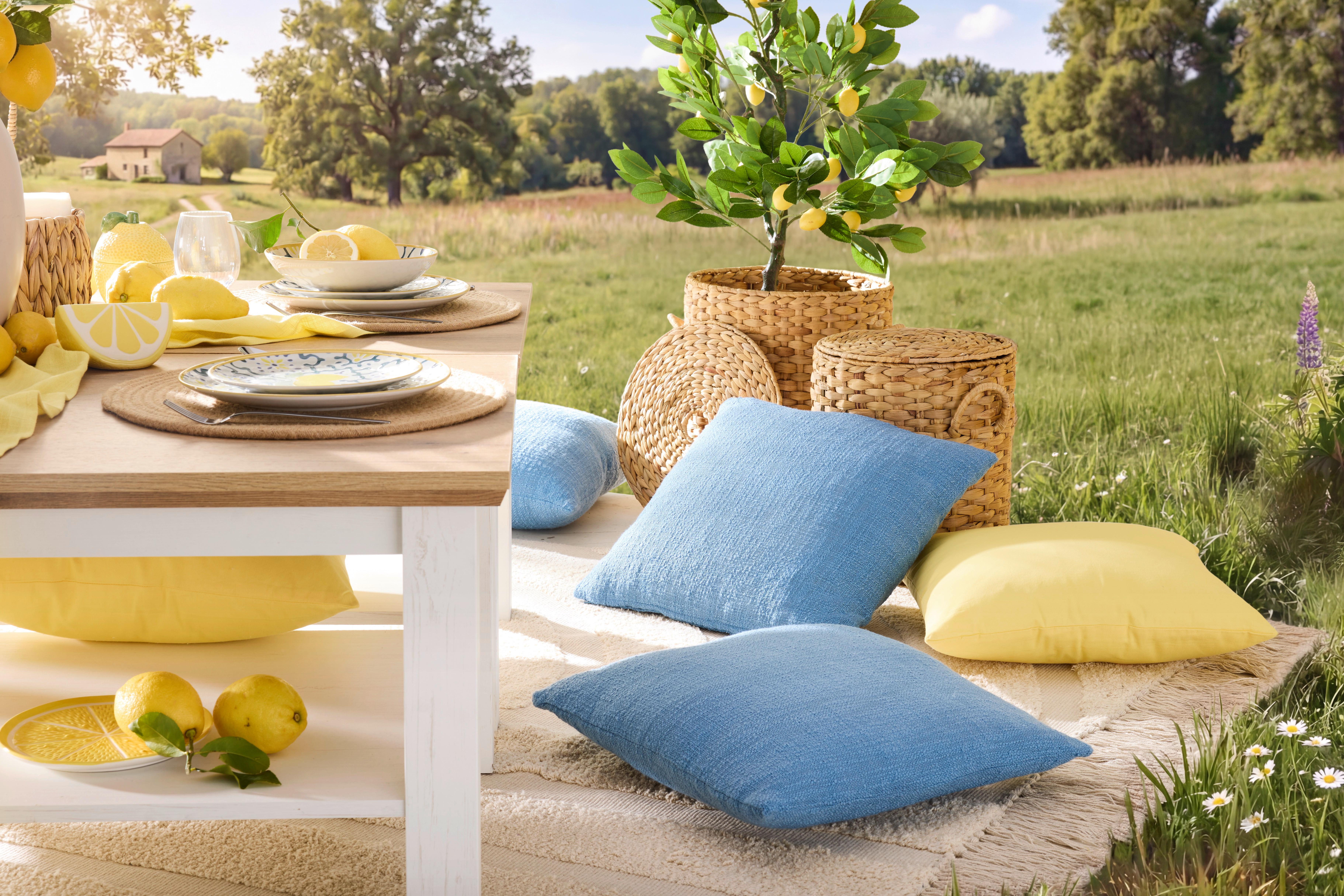 Modré a žluté dekorační polštáře Poppy a Sasha na zahradním koberci u prostřeného stolu s citrónovým servisem, proutěnými košíky a citroníku v pozadí.