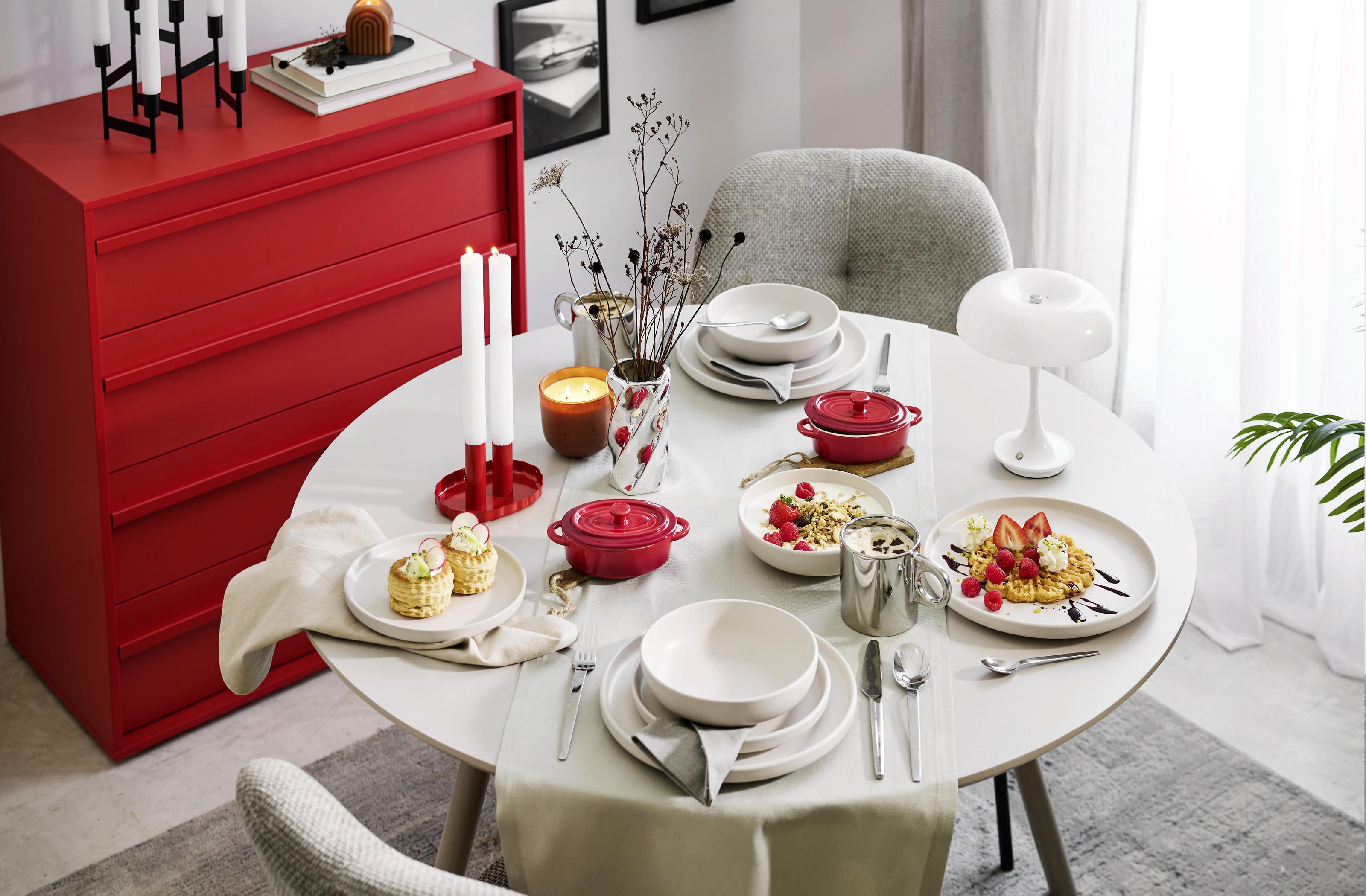 Štýlovo prestretý jedálenský stôl v retro štýle s červenými mini zapekacími miskami, bielym riadom, sviečkami a dezertmi doplnený o výraznú červenú komodu v modernom interiéri.