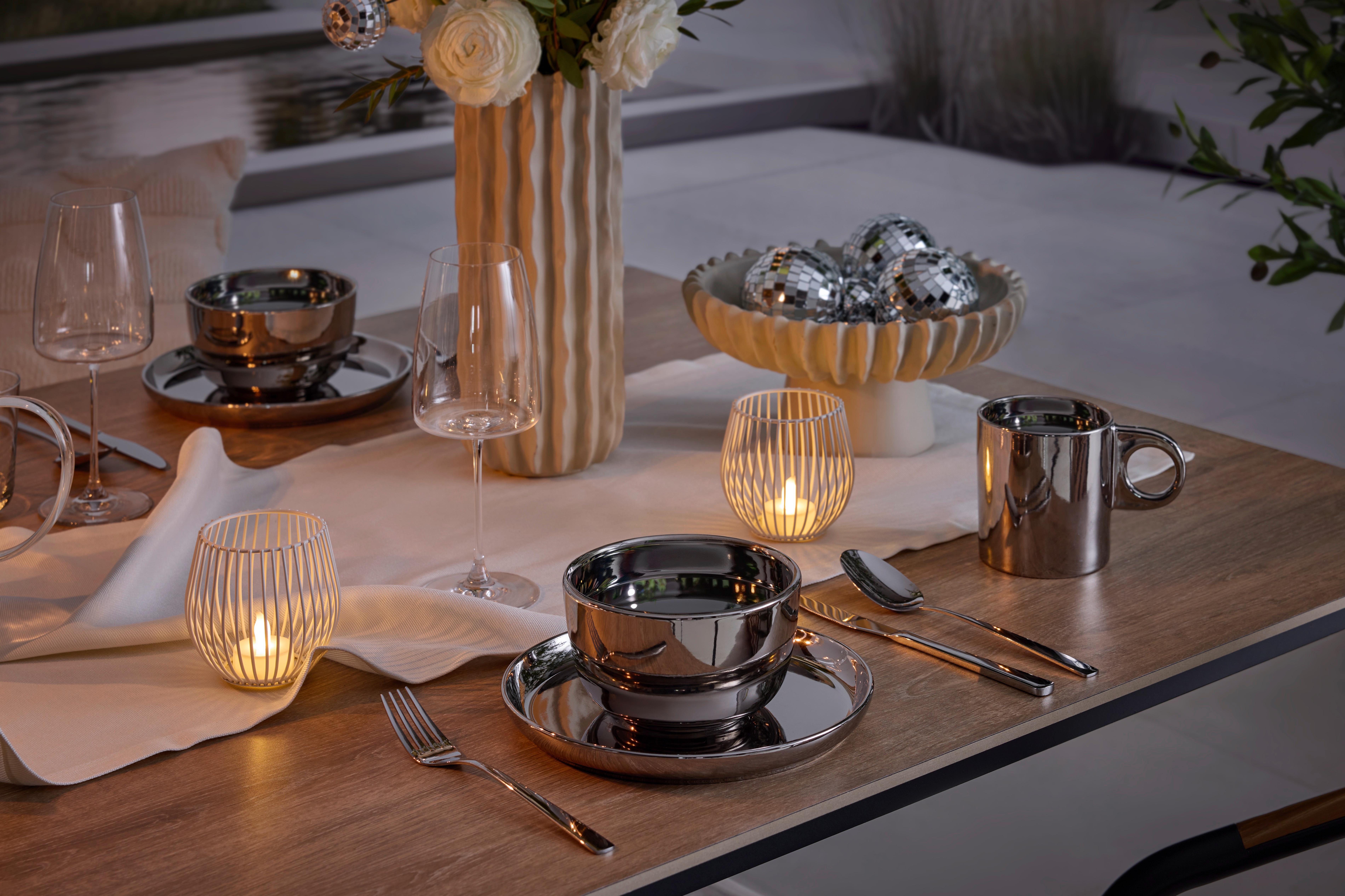 Prestretý stôl s kovovým riadom, svietnikmi, vázou s kvetmi a miskou so zrkadlovými guľami.