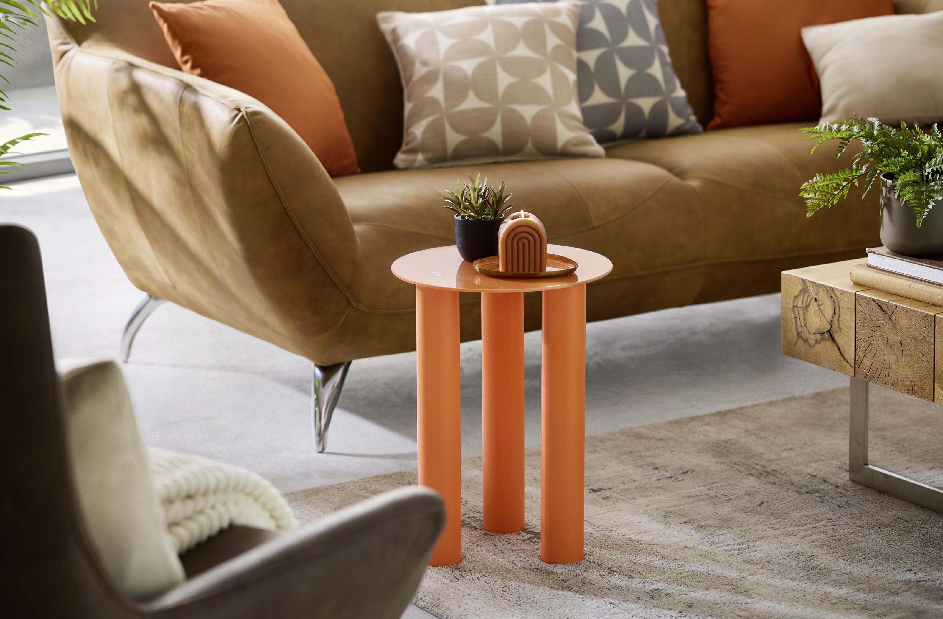 Moderný retro stolík v oranžovej farbe doplnený dekoráciou a rastlinou v obývačke s hnedou sedačkou a vzorovanými vankúšmi ktoré vytvárajú štýlový a útulný priestor.