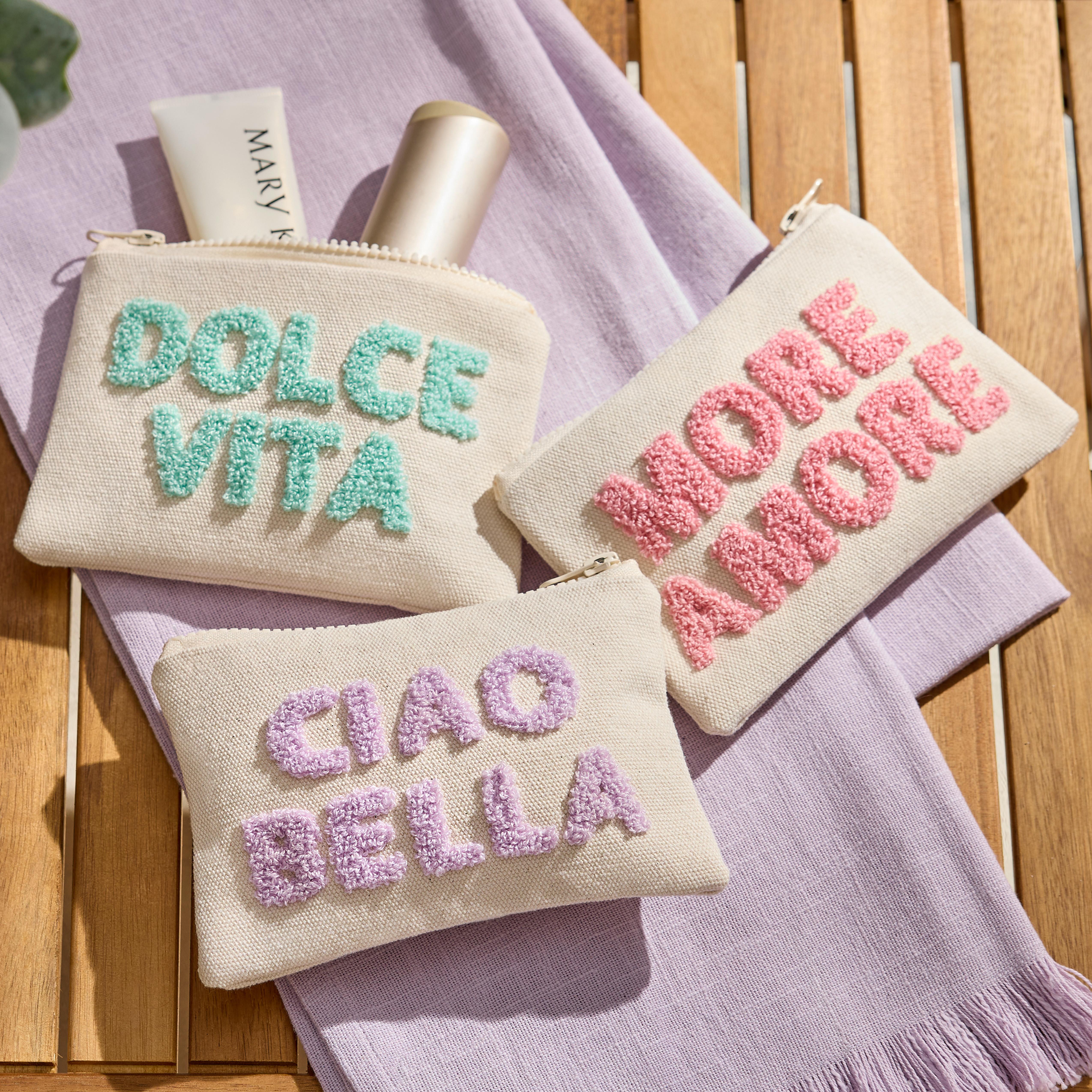 Tri textilné kozmetické taštičky s nápismi Dolce Vita, Ciao Bella a More Amore položené na fialovej osuške na drevenom stole.