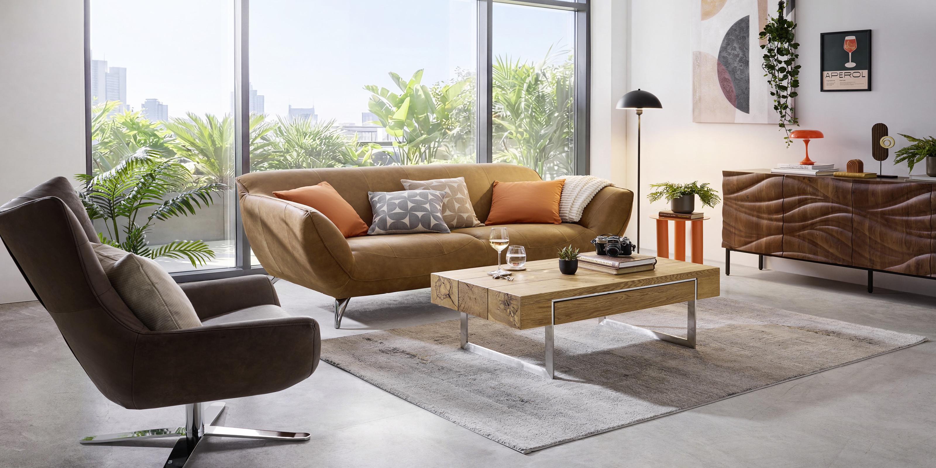 Moderná obývačka s hnedou sedačkou, kreslom, dreveným konferenčným stolíkom a retro komodou v svetlom interiéri s veľkými oknami a zeleňou.